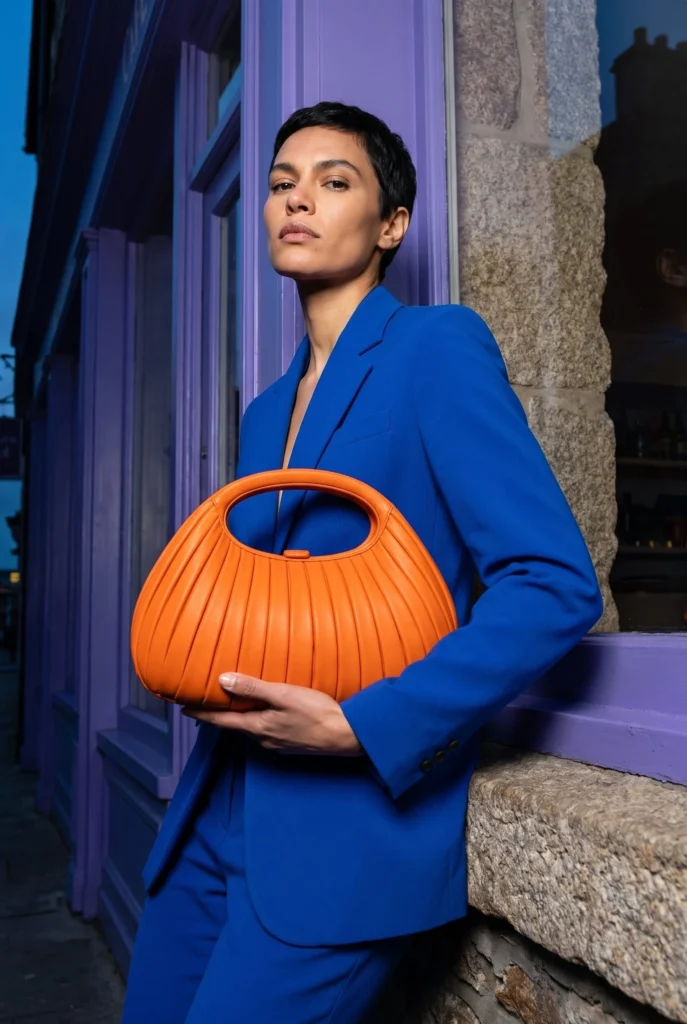 Short-haired woman in a cobalt suit supporting an orange pleated clutch outside a violet cafe, generated with Fotogenic AI