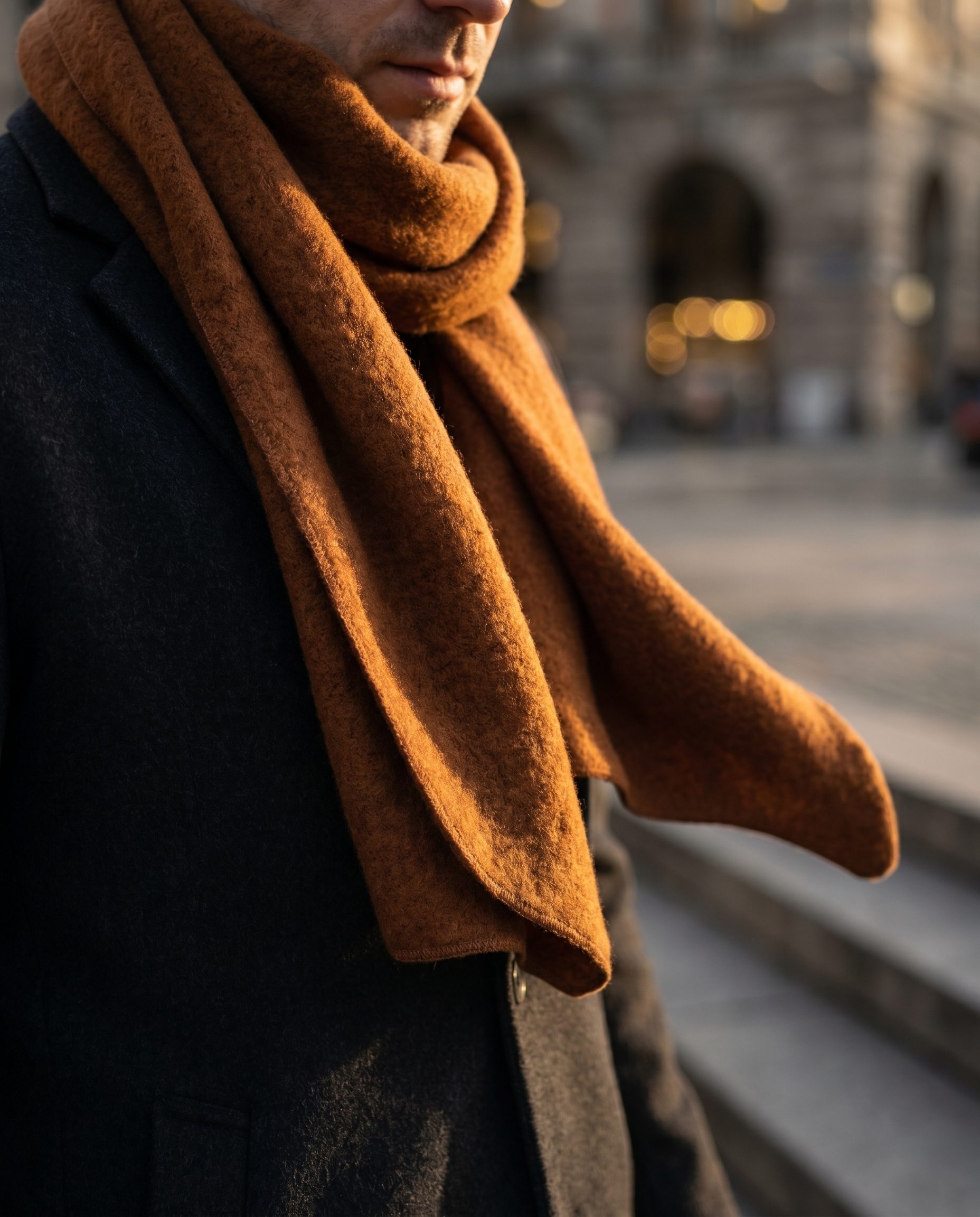 Close-up of a rust scarf styled with a dark coat in warm city light.