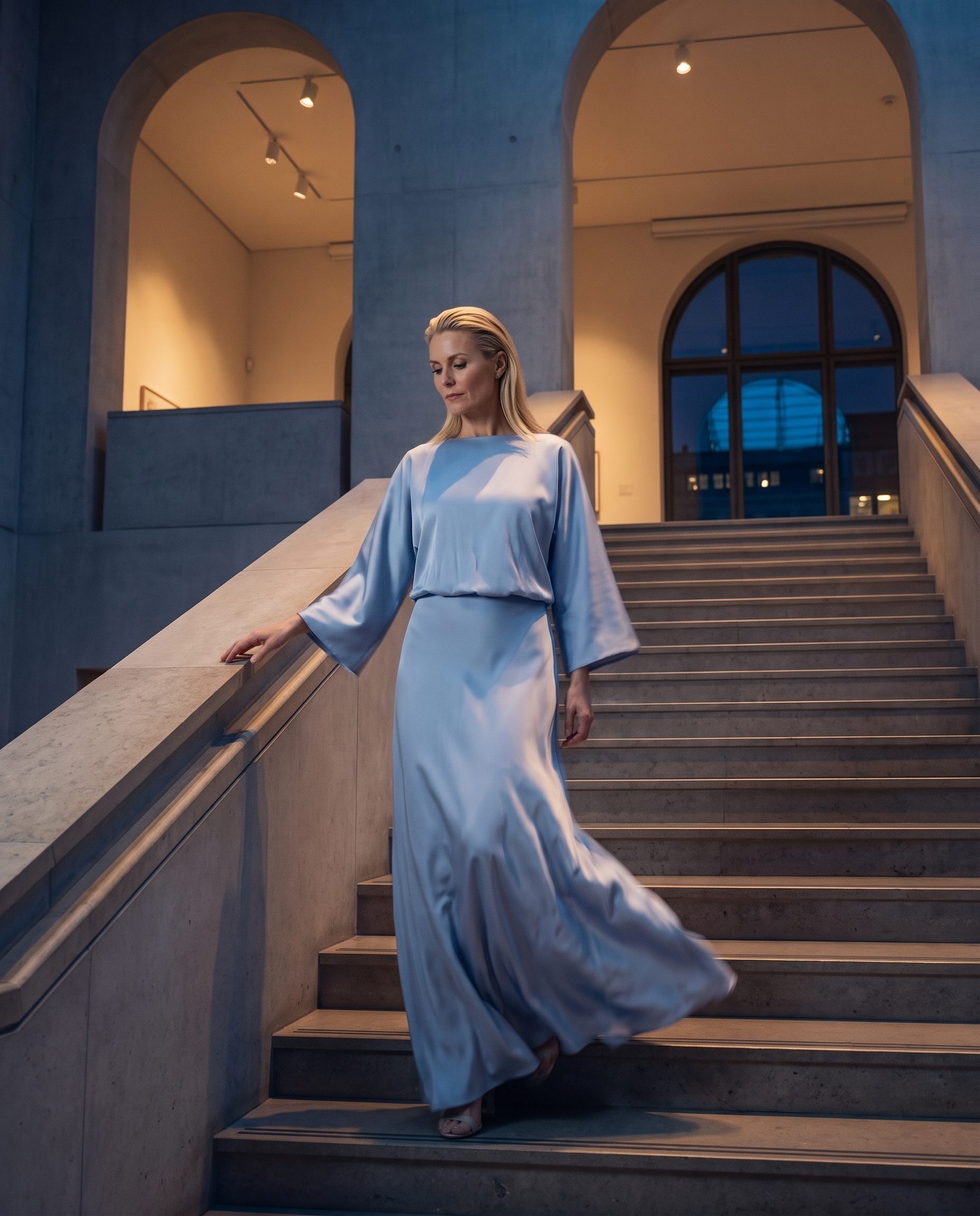 Pale blue dress worn by a blonde model on gallery stairs at blue hour.