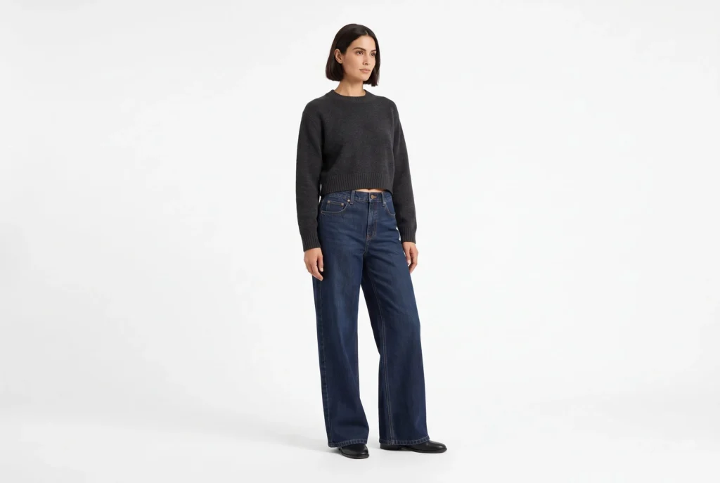 Frau in dunkelindigofarbenen, weit geschnittenen Jeans und einem anthrazitfarbenen Strickpullover in Dreiviertel-Studio-Pose.