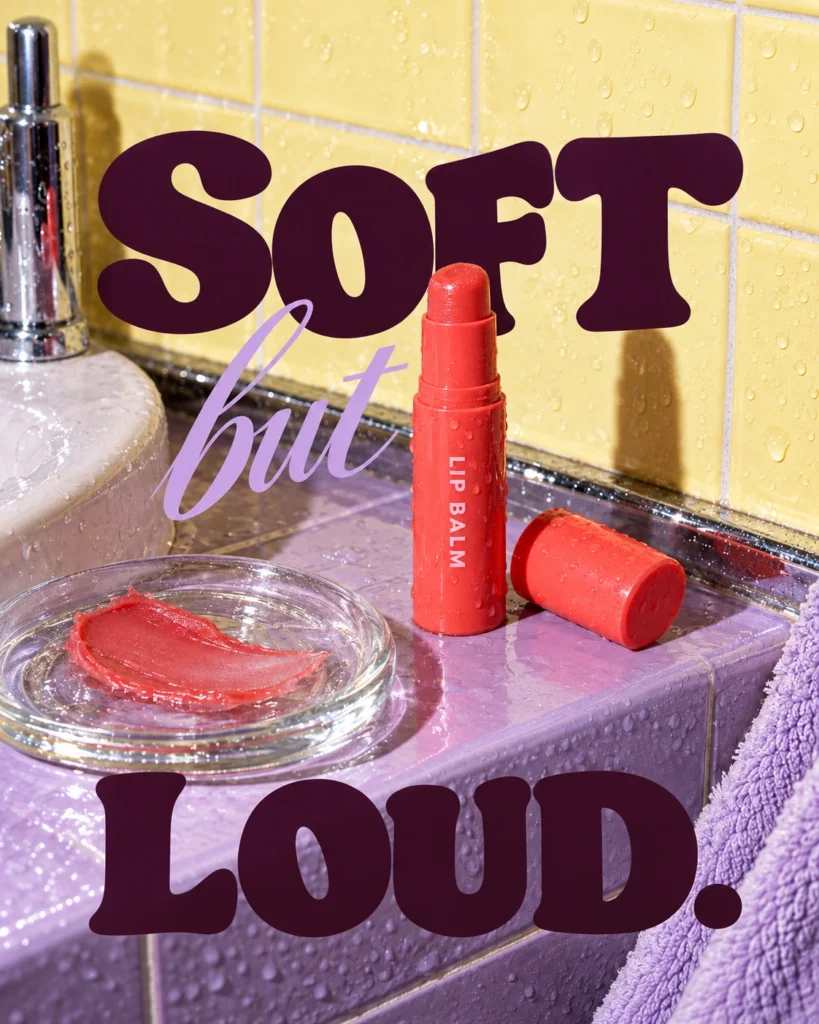 A coral lip balm tube and cap on a wet lavender ledge against yellow tiles, with bold plum Soft but Loud lettering.
