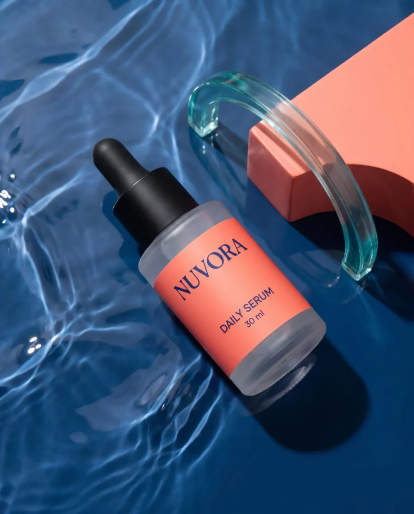 NUVORA coral-label serum with aqua glass and coral shapes on a midnight-blue surface