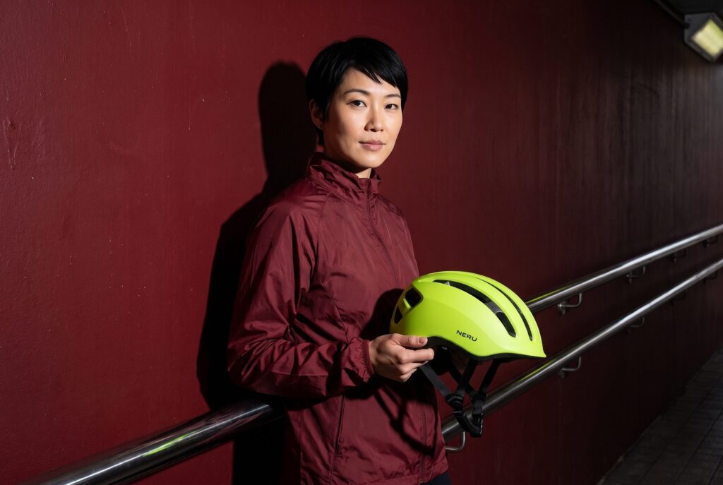 Woman holding a lime-green NERU cycling helmet beside a burgundy wall.
